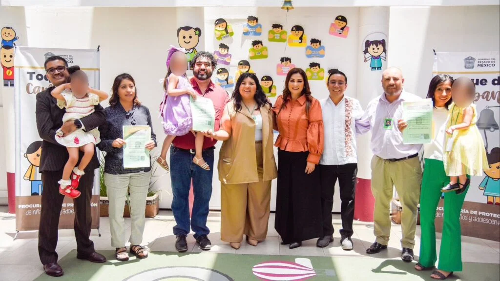 Grupo de personas posando en un espacio institucional infantil, varias sostienen documentos oficiales y dos niñas son cargadas por personas adultas; al fondo se observan decoraciones coloridas y señalética gubernamental.