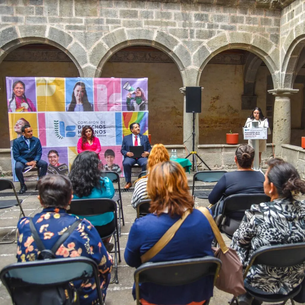 Evento en patio con arquitectura de arcos de piedra donde varias personas están sentadas frente a un escenario. En el fondo hay un banner de la Comisión de Derechos Humanos del Estado de México con imágenes de distintas personas y colores brillantes. A la derecha, una persona está de pie en un atril dando una intervención, mientras tres personas permanecen sentadas como panelistas.
