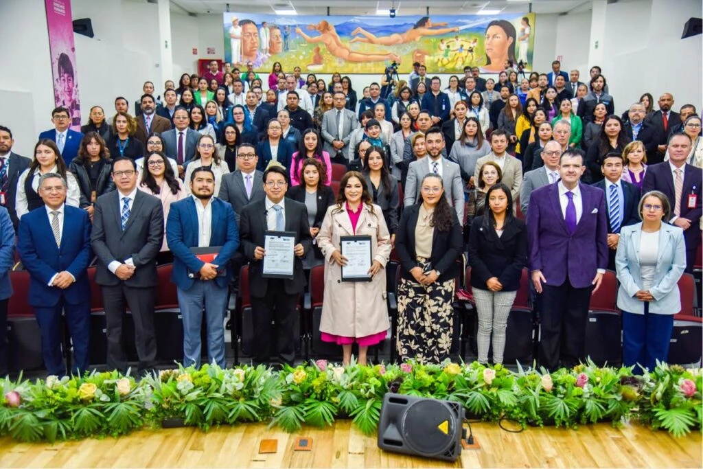 Fotografía grupal en auditorio con numerosas personas de pie en varias filas; al frente, varias personas sostienen documentos enmarcados sobre un escenario con arreglos florales.