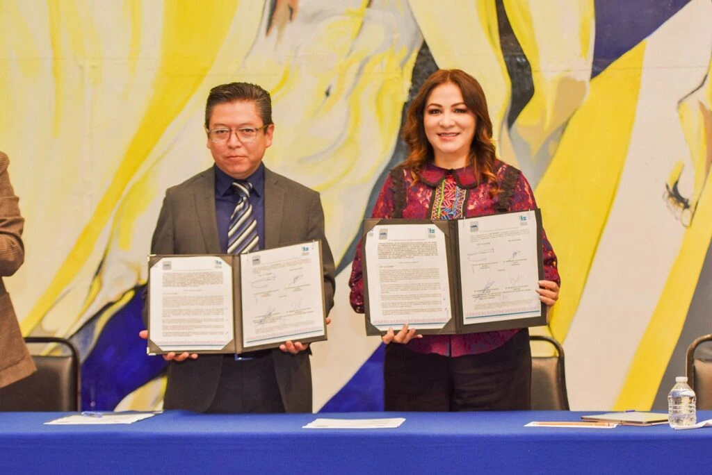 Dos personas de pie sostienen carpetas abiertas mostrando documentos firmados durante una ceremonia formal, con un mural artístico de fondo.