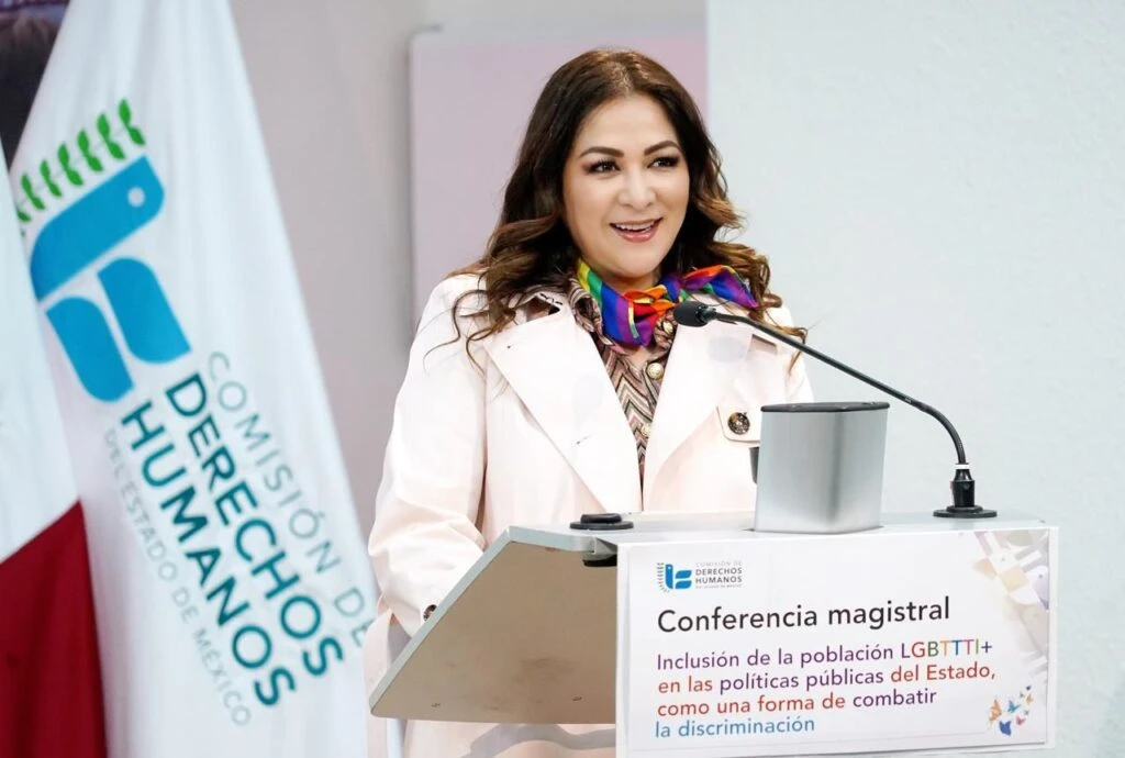 Persona hablando en un podio durante una conferencia magistral, con un letrero que menciona inclusión de la población LGBTTTI+ en políticas públicas y un emblema institucional al fondo.