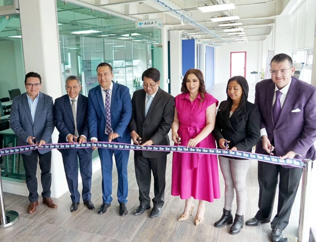 Ceremonia de corte de listón con varias personas alineadas sosteniendo tijeras frente a una cinta institucional, en un pasillo con aulas de cristal y señalización visible.