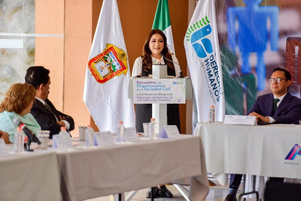 Persona hablando desde un podio en un evento sobre organizaciones de la sociedad civil y derechos humanos, con banderas institucionales a los lados y asistentes sentados en mesas al frente.