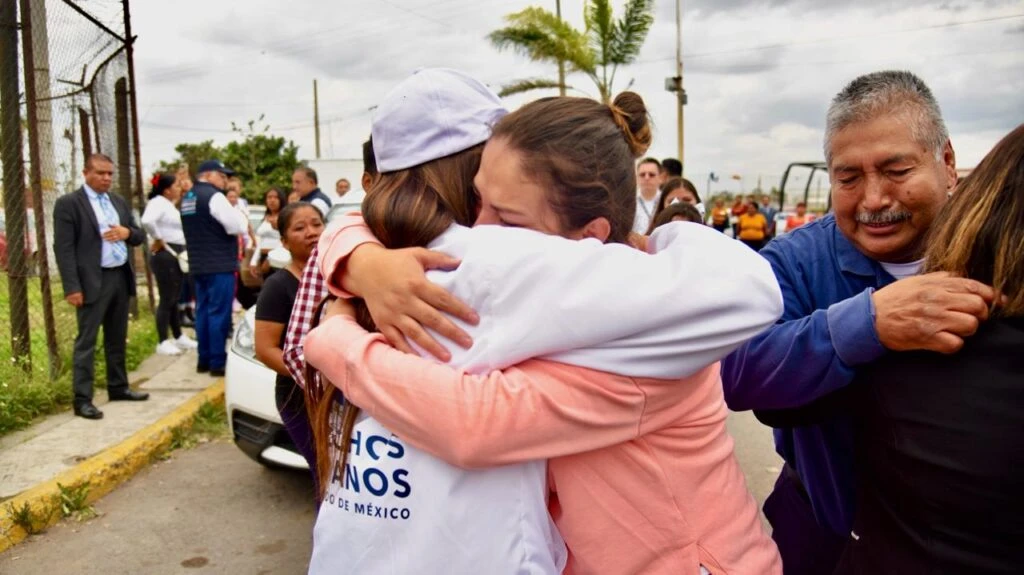 Dos personas se abrazan en una calle junto a una cerca metálica mientras otras personas permanecen alrededor observando; se aprecian vehículos estacionados y un entorno urbano abierto.