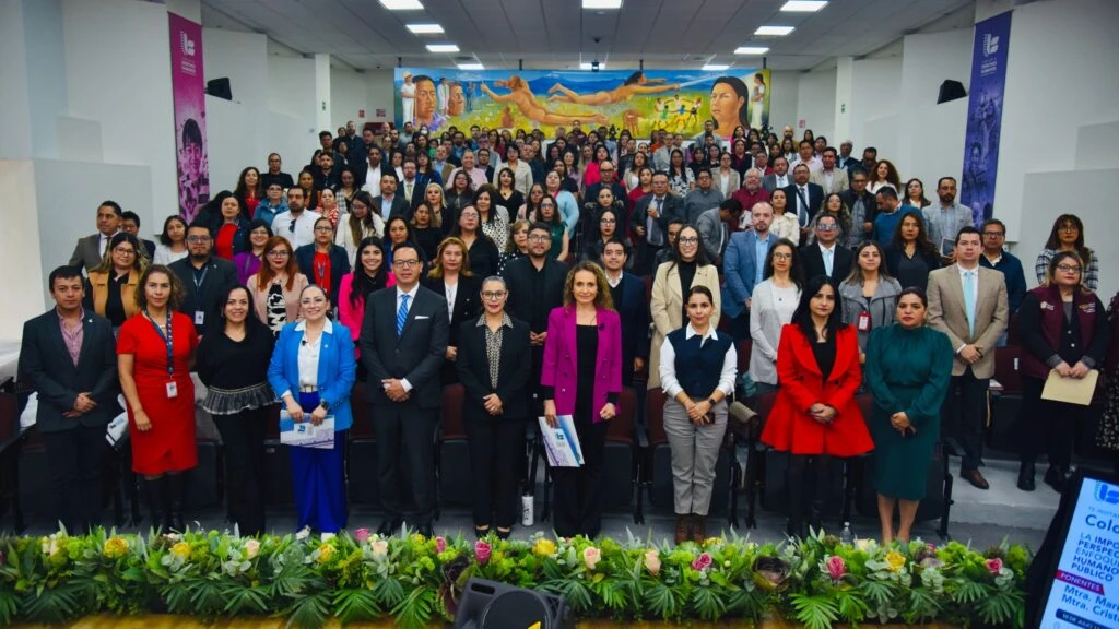 Fotografía grupal en auditorio con numerosas personas organizadas en filas sobre las gradas; al frente hay una fila principal con personas de pie frente al escenario decorado con arreglos florales y un mural colorido al fondo.