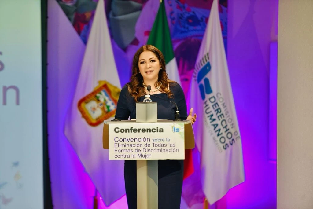 Persona dando una conferencia desde un podio con micrófono, frente a banderas institucionales y un fondo iluminado en tonos morados; el atril muestra el texto sobre la Convención para la Eliminación de Todas las Formas de Discriminación contra la Mujer.