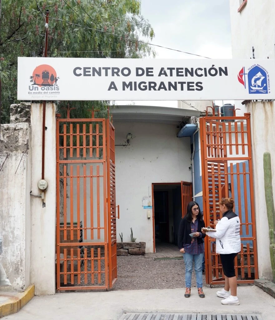 Fachada de un centro con letrero que dice “Centro de Atención a Migrantes”, con una reja naranja abierta mientras dos personas intercambian documentos en la entrada.