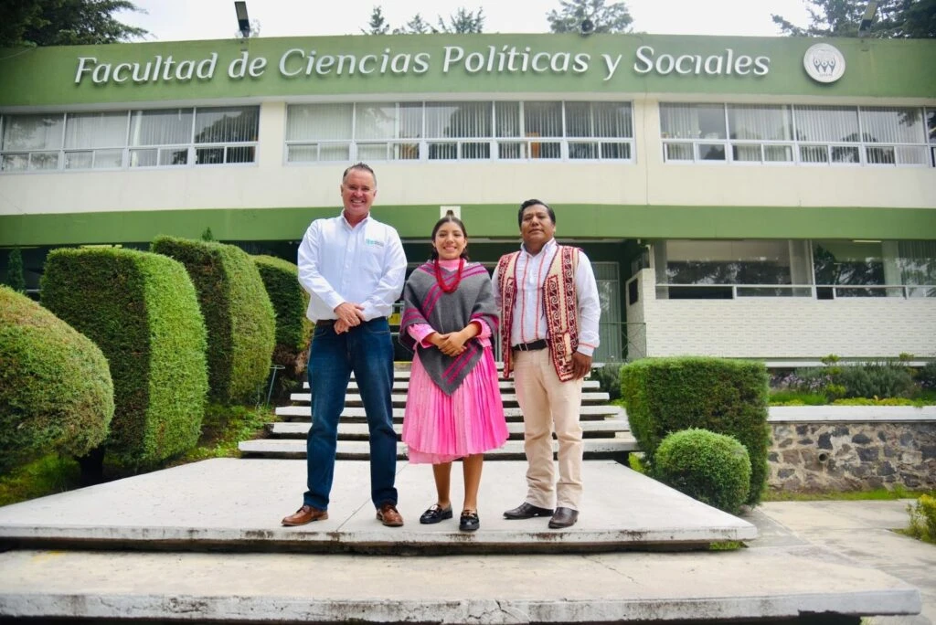 Tres personas posando de pie frente a un edificio que muestra el rótulo “Facultad de Ciencias Políticas y Sociales”, con áreas verdes y escaleras en primer plano.