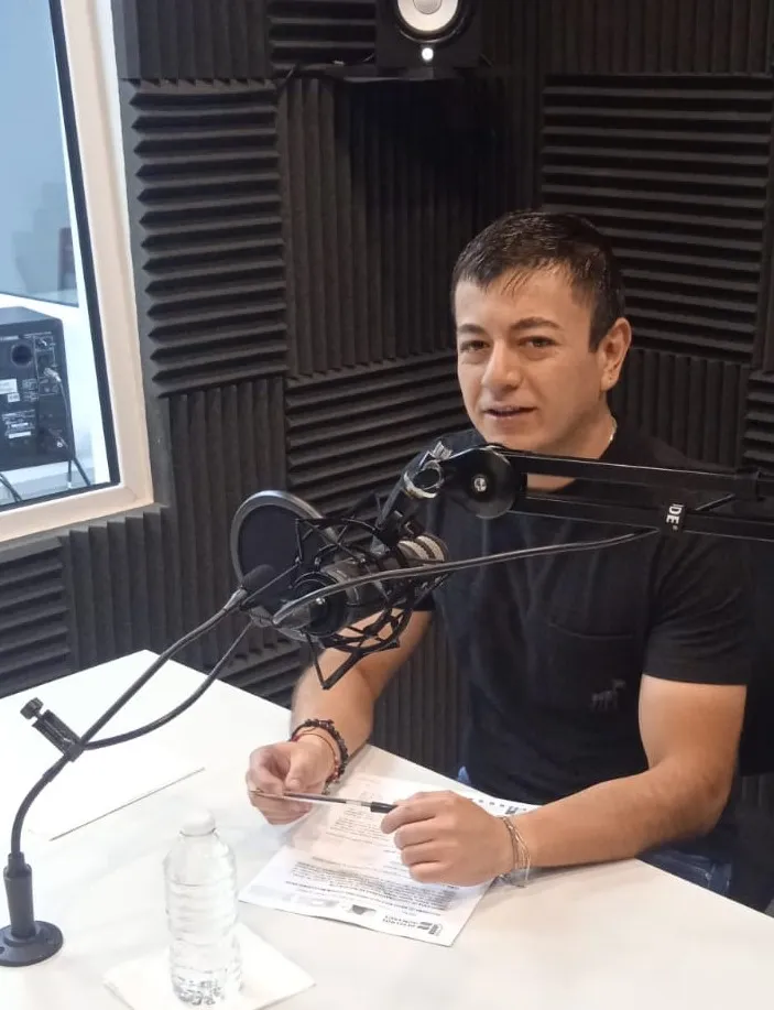 Persona sentada frente a un micrófono profesional en un estudio de grabación con paneles acústicos negros en las paredes. Sobre la mesa hay una hoja con texto, un bolígrafo y una botella de agua. La persona está hablando mientras sostiene el bolígrafo y se encuentra frente a un brazo articulado con micrófono y filtro antipop.