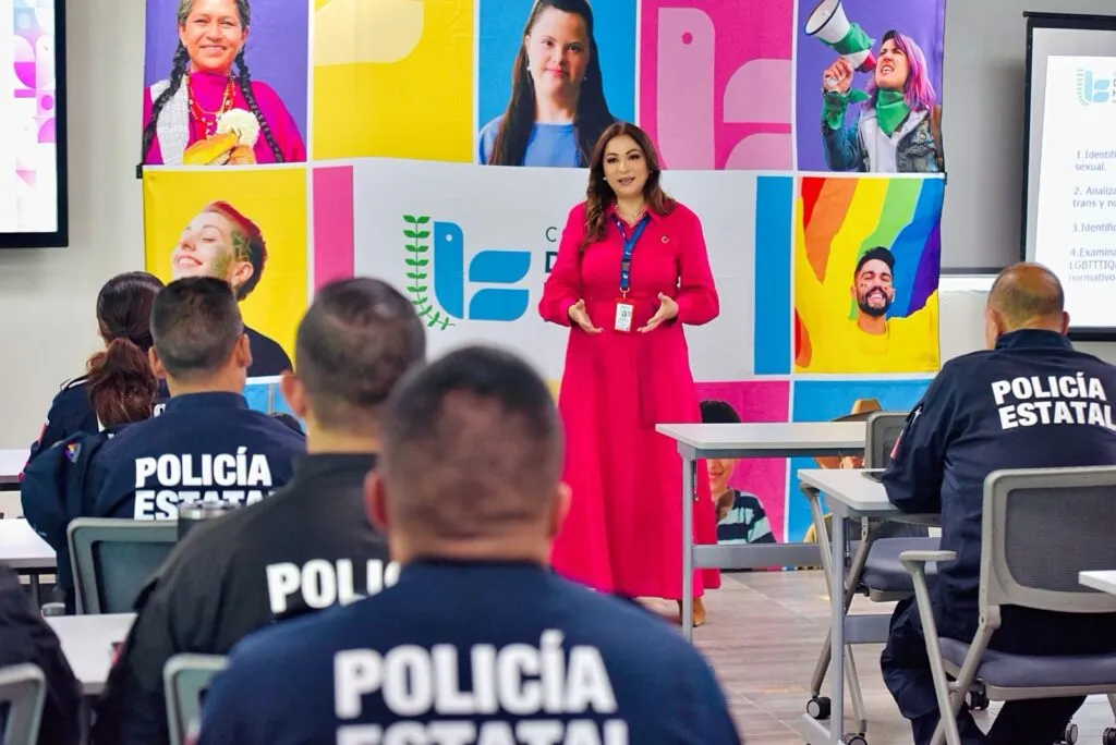 Persona impartiendo una capacitación al frente de un salón a elementos de policía estatal sentados en filas. En el fondo se observa un mural colorido con retratos y una pantalla de presentación; en el uniforme de los asistentes se lee “POLICÍA ESTATAL”.
