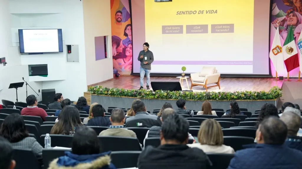 Vista frontal de un auditorio donde una persona expone en un escenario frente a una audiencia sentada. En la pantalla se proyecta una presentación titulada “Sentido de vida” con varias preguntas, y a un lado se encuentra la bandera de México y mobiliario de escenario.