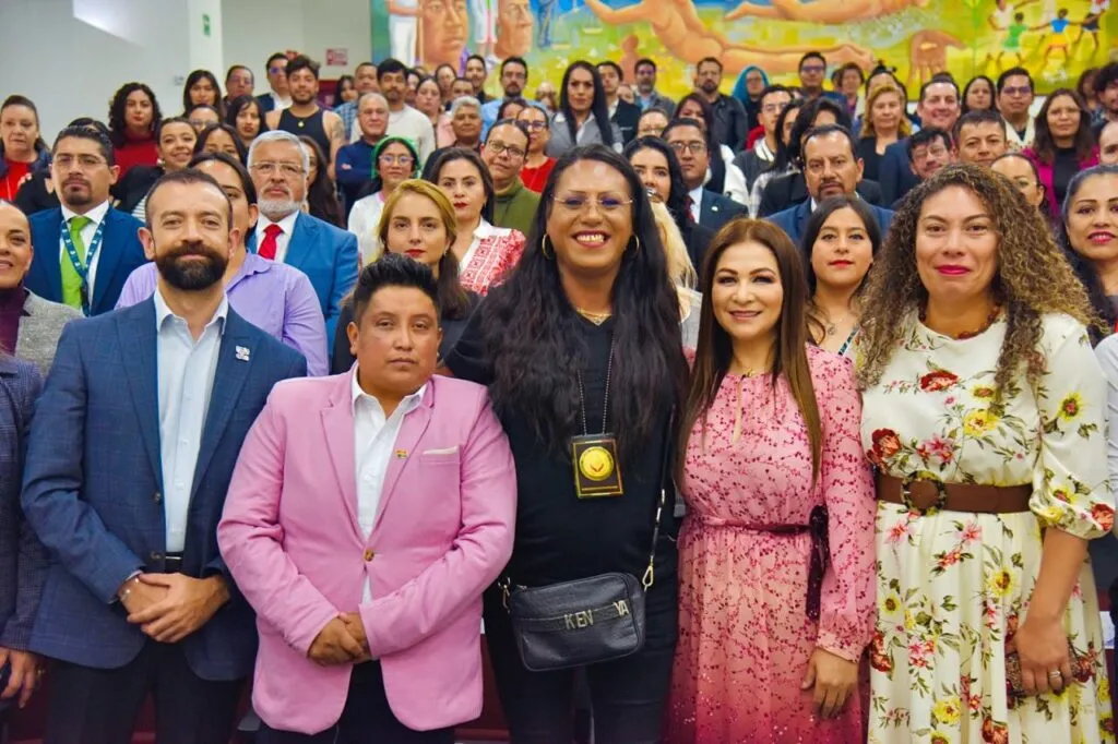 Grupo numeroso de personas reunidas en un auditorio para una fotografía grupal. En primer plano hay varias personas de pie alineadas, mientras al fondo se observan más asistentes en gradas, junto con un mural colorido en la pared.