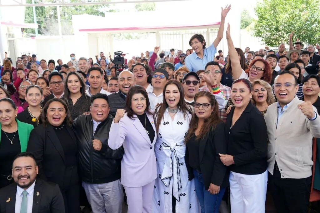 Grupo numeroso de personas reunidas en un evento público, muchas de ellas al frente levantan las manos en señal de celebración mientras posan para una fotografía. En el fondo se observa una multitud y un espacio abierto con estructura techada.