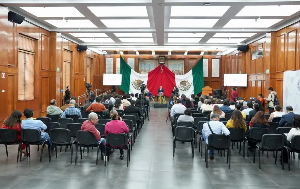 Vista general de un auditorio con filas de sillas ocupadas por asistentes que observan hacia un escenario al frente. En el escenario hay una mesa con personas participantes, equipo de grabación y una gran bandera de México colocada como fondo en un espacio con paredes de madera.