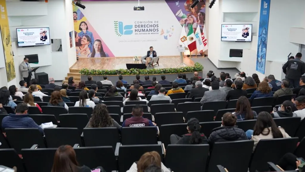 Vista amplia de un auditorio moderno con un escenario donde una persona participa sentada al centro, frente a una audiencia numerosa. Al fondo se observa el logotipo de la Comisión de Derechos Humanos del Estado de México, junto con pantallas laterales y banderas.