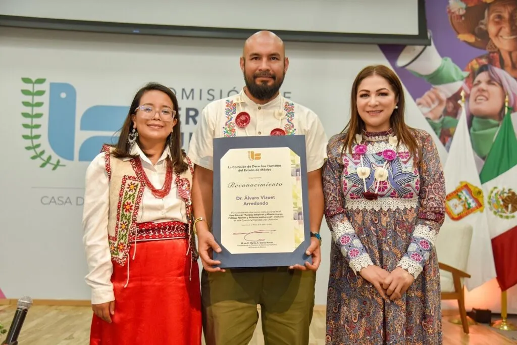 Tres personas posan en un escenario institucional sosteniendo un reconocimiento enmarcado al centro. El fondo muestra el logotipo de la Comisión de Derechos Humanos del Estado de México y banderas oficiales; las personas visten indumentaria con bordados tradicionales.