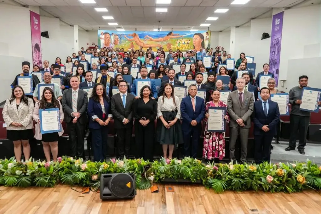Fotografía grupal de decenas de personas en un auditorio, organizadas en varias filas y sosteniendo reconocimientos enmarcados. En la parte frontal hay personas de pie en primera fila y decoración con plantas, mientras al fondo se observa un mural colorido.