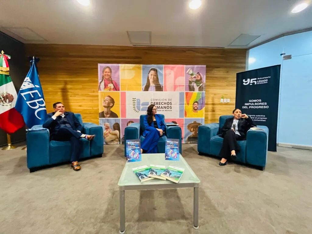 Panel de discusión en un espacio interior con paredes de madera y fondo institucional de la Comisión de Derechos Humanos del Estado de México. Tres personas se encuentran sentadas en sillones individuales sobre un escenario, con una mesa de centro que contiene publicaciones, mientras a un lado se observan banderas oficiales y un banner conmemorativo.