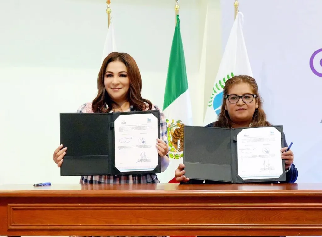 Dos personas posan detrás de una mesa mostrando carpetas abiertas con documentos firmados, en una ceremonia institucional. Al fondo se observan banderas y un espacio oficial donde se realiza la firma de convenio.