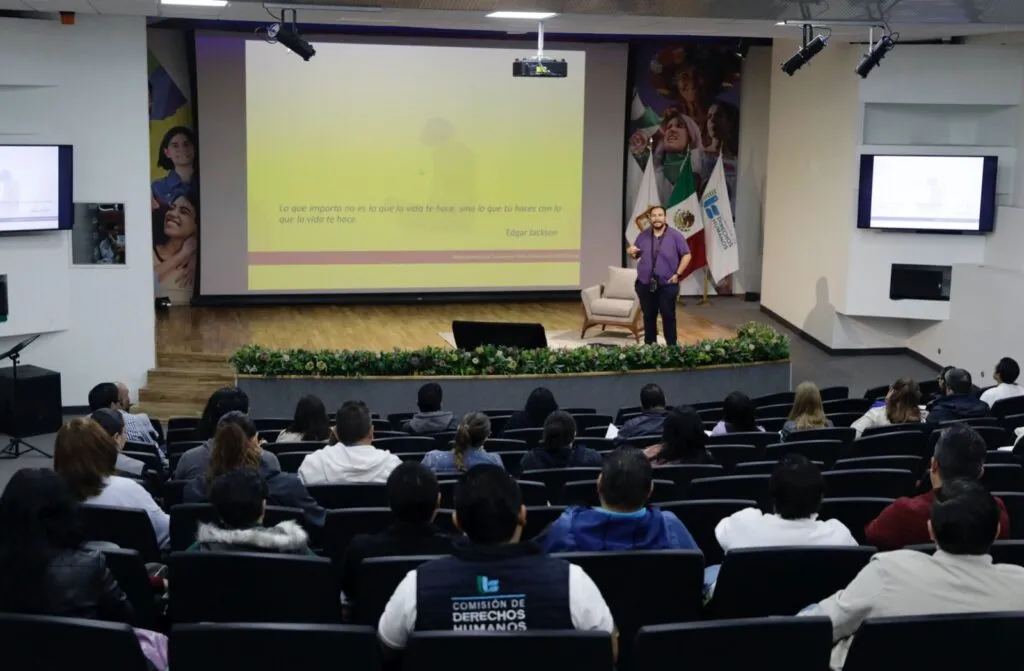 Primer plano de una conferencia donde una persona presenta frente a una pantalla con texto que incluye temas como realidad, sentimientos, adaptación y reinvención. El expositor está de pie sobre el escenario con apoyo visual, y el público se encuentra sentado escuchando.