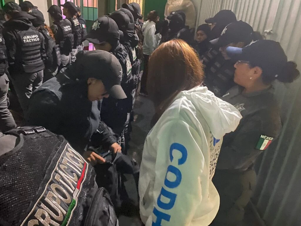 Personal de seguridad con equipo táctico y observadores de la Comisión de Derechos Humanos en una revisión dentro de un espacio cerrado con iluminación artificial.