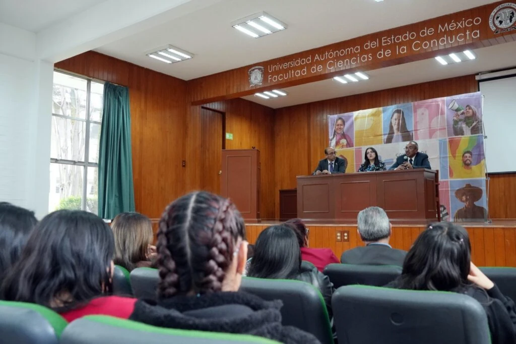 Auditorio universitario con asistentes sentados frente a un estrado donde tres personas participan en una mesa de diálogo; al fondo se observa el letrero de la Facultad de Ciencias de la Conducta de la UAEMéx.
