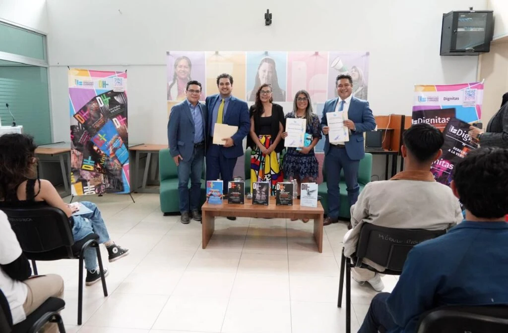 Grupo de participantes en la parte frontal de una sala sosteniendo reconocimientos durante un evento de presentación; al centro hay una mesa con publicaciones y al fondo banners de derechos humanos.