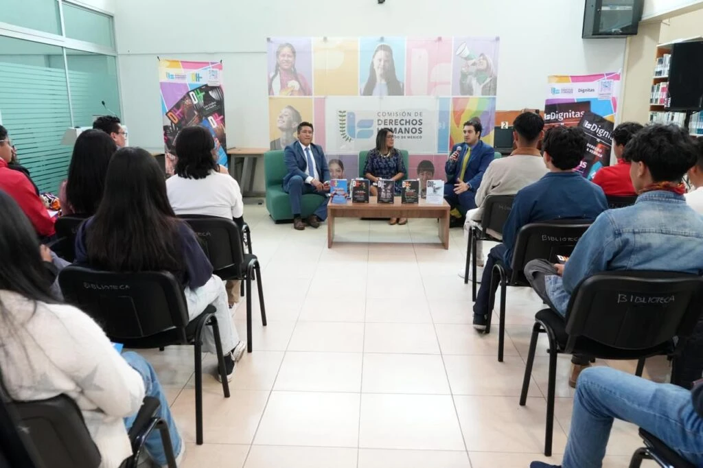 Audiencia sentada en filas frente a un panel de personas en una mesa, durante una charla o presentación con material editorial exhibido al centro.