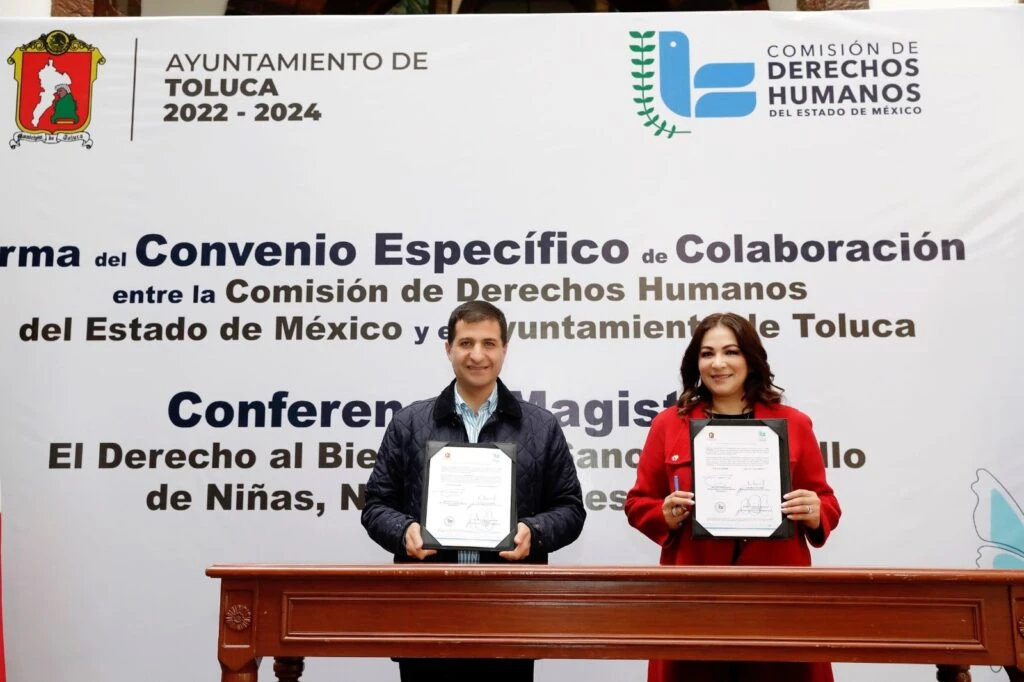 Dos personas posan detrás de una mesa mostrando documentos firmados durante la firma de un convenio de colaboración entre el Ayuntamiento de Toluca y la Comisión de Derechos Humanos del Estado de México, frente a una lona oficial.