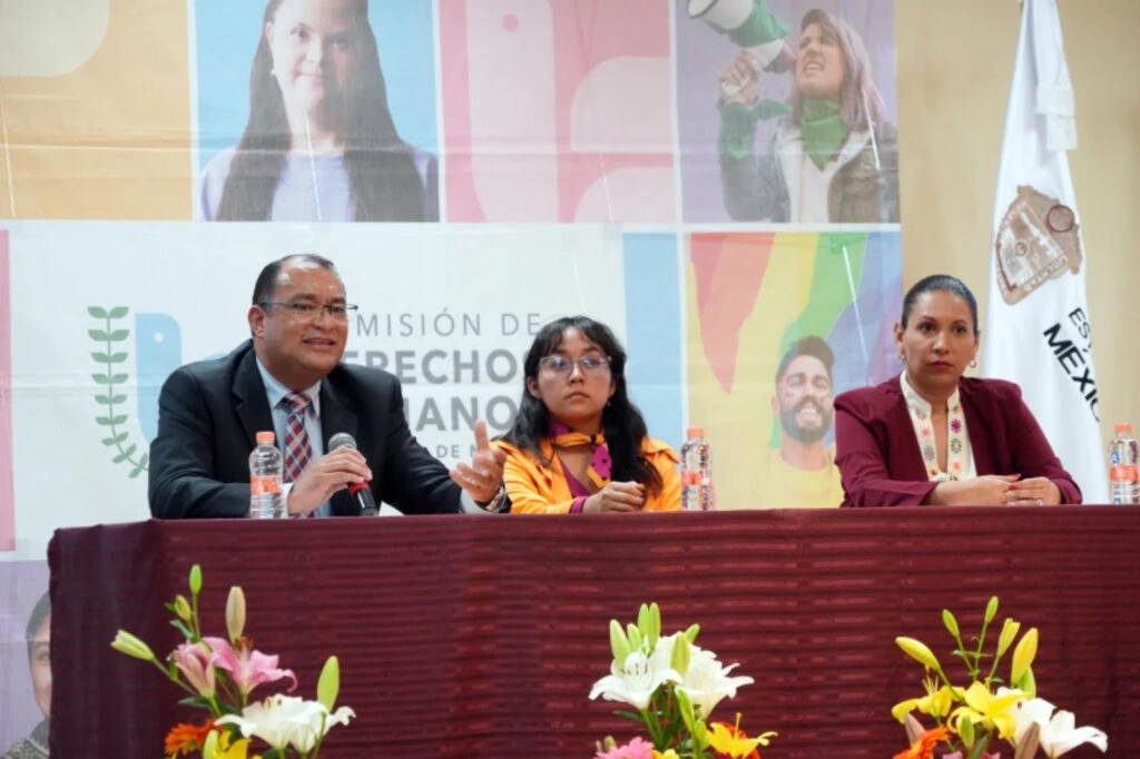 Mesa de panel con tres personas sentadas frente a micrófonos y botellas de agua durante una presentación institucional; al fondo se observa un cartel con el logotipo de la Comisión de Derechos Humanos y una composición gráfica con rostros y colores.