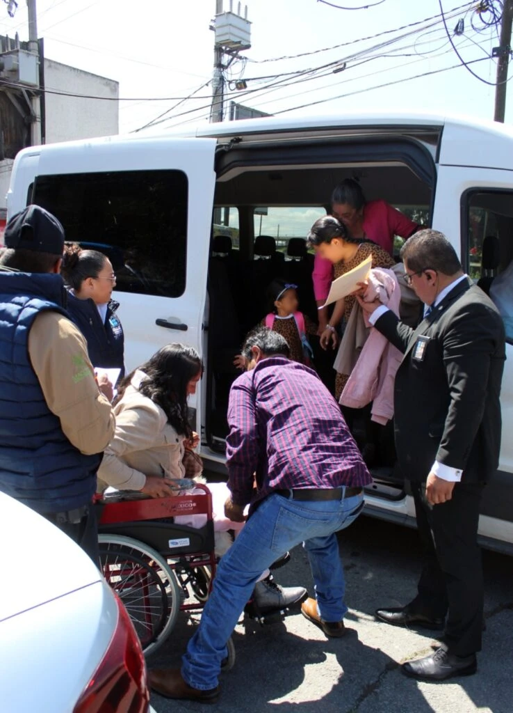 Varias personas se agrupan alrededor de una camioneta blanca con la puerta lateral abierta en la vía pública; algunas ayudan a descender a una persona en silla de ruedas, mientras otras sostienen documentos y prendas, con cables y construcciones visibles en el fondo urbano.
