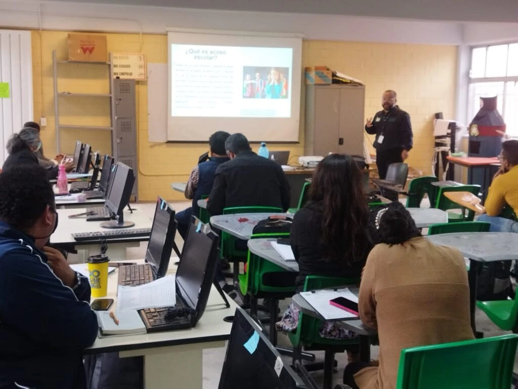 Aula de informática con varias personas sentadas frente a computadoras mientras una persona expone al frente junto a una proyección sobre una pantalla que muestra información sobre acoso escolar.