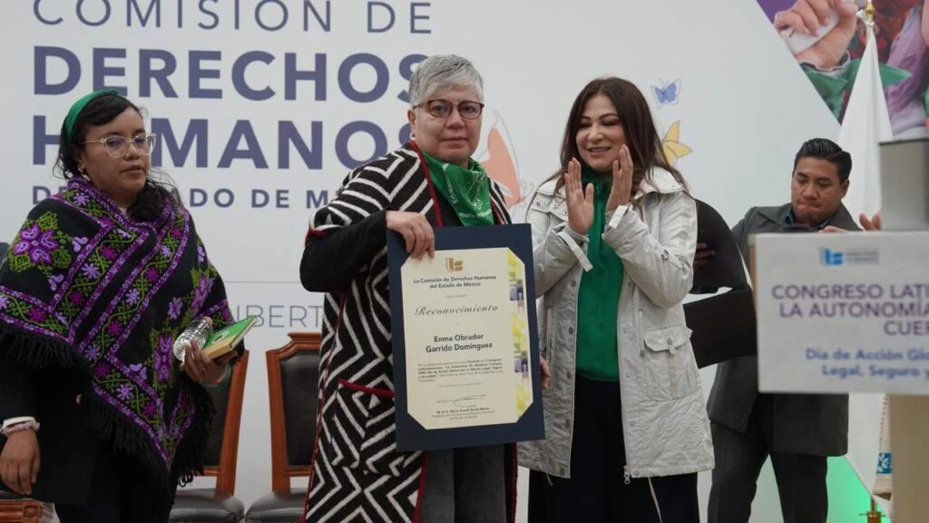 Persona en un podio durante un evento institucional levanta un pañuelo verde, con banderas oficiales detrás y un letrero que menciona un congreso latinoamericano sobre la autonomía y derechos.