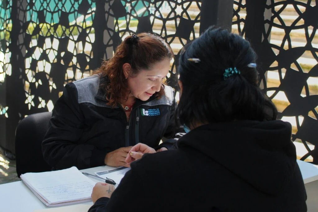 Dos personas sentadas frente a frente en una mesa durante una atención personalizada; una de ellas revisa documentos y toma notas en un cuaderno mientras dialogan, en un espacio con panel decorativo al fondo.