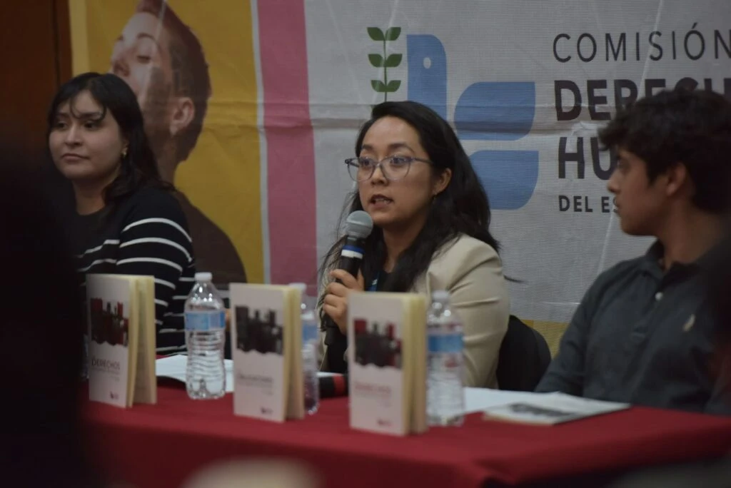 Persona hablando con micrófono en un panel, sentada junto a otras personas en una mesa con libros exhibidos y botellas de agua, frente a un fondo con el logotipo de la Comisión de Derechos Humanos del Estado de México.