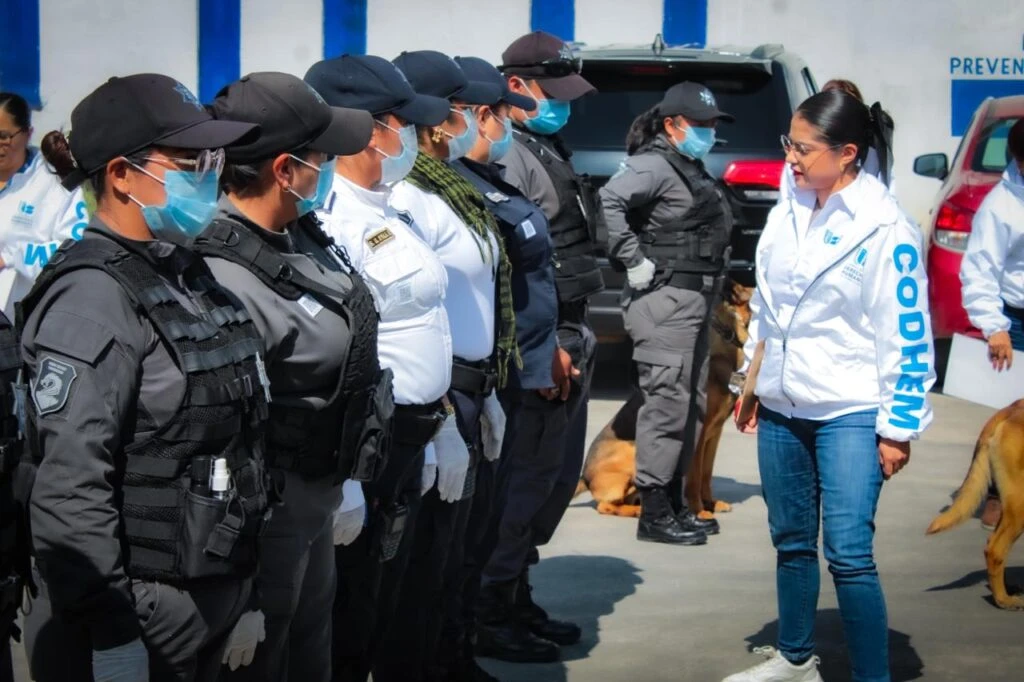 Formación de personal uniformado alineado en fila durante una supervisión en exteriores; al frente, una persona con chamarra institucional dialoga con el grupo, y se observan vehículos y un perro de trabajo al fondo.