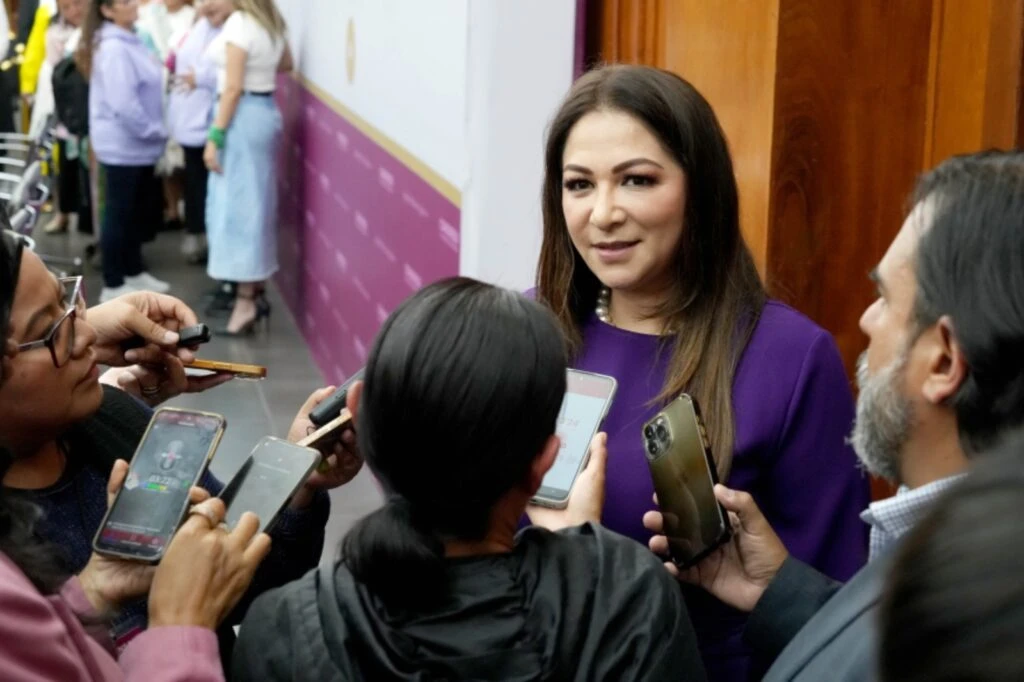 Persona vestida con ropa de color púrpura responde a varias preguntas de reporteros, mientras múltiples dispositivos móviles y grabadoras se acercan en un pasillo interior con otras personas al fondo.