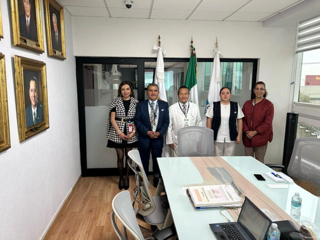 Fotografía tomada en una sala de juntas con piso de madera y paredes blancas. En el centro aparecen cinco personas de pie frente a una mesa de reuniones; dos personas visten ropa formal, una persona usa bata blanca y otras dos llevan vestimenta institucional.