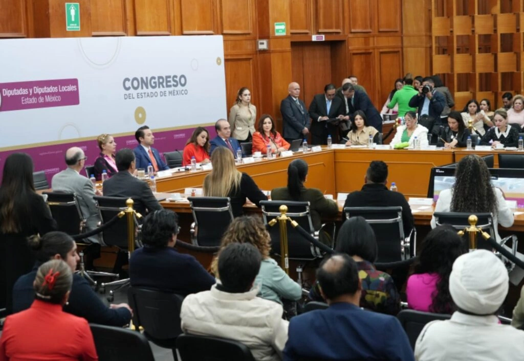 Sesión de trabajo en una sala del Congreso del Estado de México con paredes de madera. En el centro hay una mesa en forma de herradura rodeada de personas sentadas y de pie. En la parte frontal se observa un panel con el texto “Congreso del Estado de México” y “Diputadas y Diputados Locales Estado de México”. Varias personas participan mientras otras observan desde el público; al fondo hay personal de apoyo y medios de comunicación.