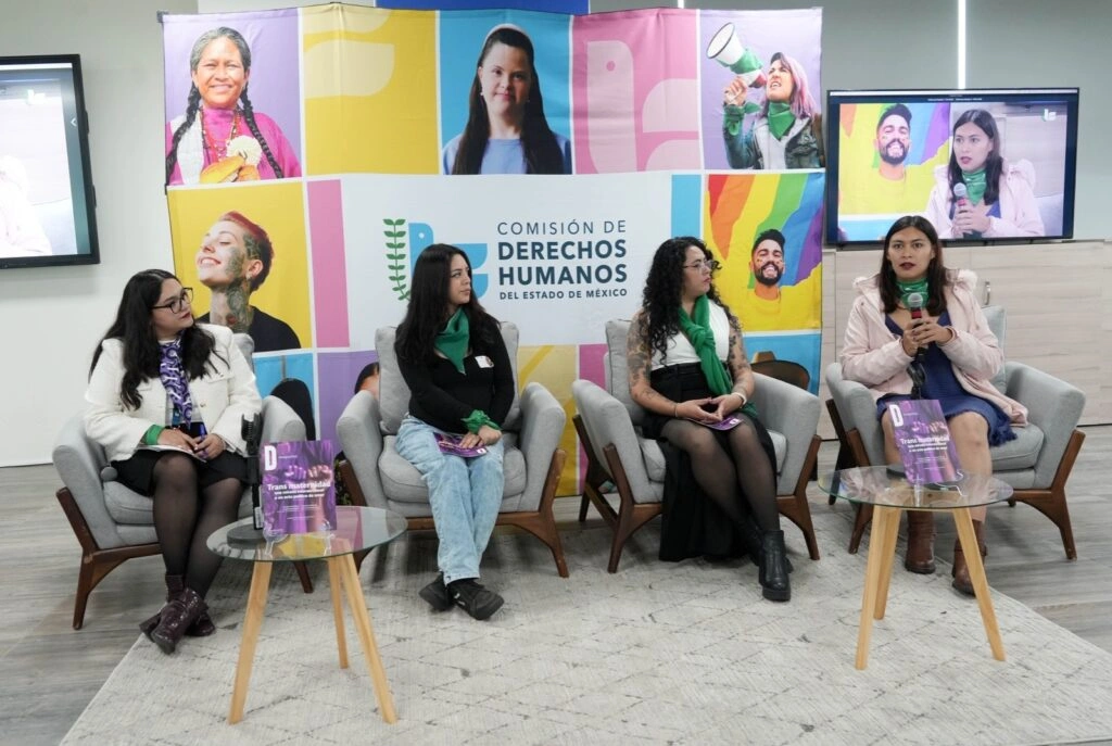 Panel de diálogo en un espacio institucional con cuatro personas sentadas en sillones individuales. Cada persona sostiene o tiene cerca una revista. Al fondo hay un mural colorido con retratos y el logotipo de la Comisión de Derechos Humanos del Estado de México. A la derecha se observa una pantalla que muestra la transmisión del evento. En el centro hay mesas pequeñas con publicaciones y objetos decorativos.
