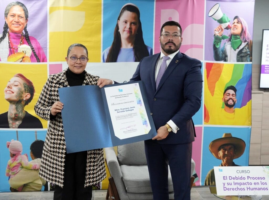 Entrega de reconocimiento en un espacio institucional. Dos personas se encuentran de pie sosteniendo un diploma abierto. Al fondo hay un mural colorido con retratos de diversas personas y el logotipo de la Comisión de Derechos Humanos del Estado de México. A un lado se observa un letrero con el título “Curso: El Debido Proceso y su Impacto en los Derechos Humanos”. También hay mobiliario como sillones y mesas.