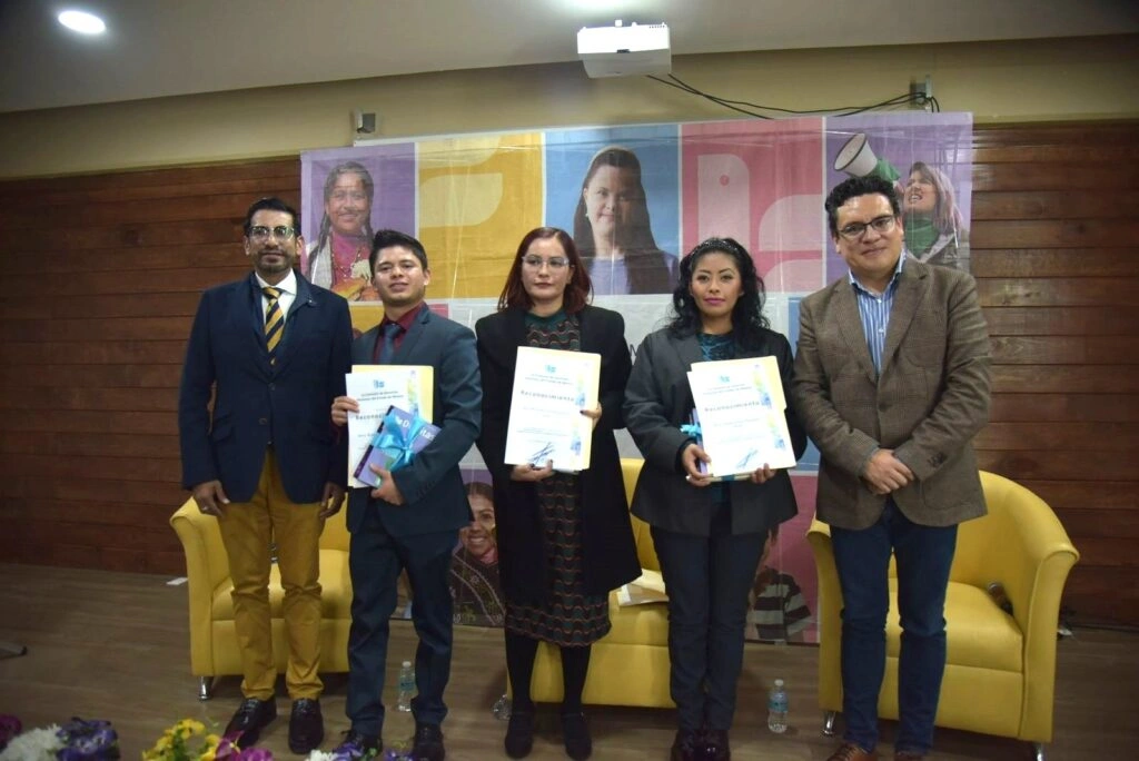 Fotografía grupal en el escenario de un evento. Cinco personas se encuentran de pie frente a un fondo institucional con imágenes y colores diversos. Tres de las personas sostienen reconocimientos o diplomas. Se observan sillones amarillos detrás y botellas de agua en el suelo frente al escenario.