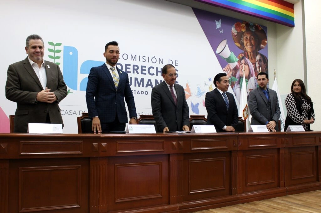 Grupo de personas de pie detrás de una mesa larga en un evento formal. Todas se encuentran alineadas frente al público, con gafetes y micrófonos sobre la mesa. En el fondo se observa un mural con ilustraciones y el logotipo de la Comisión de Derechos Humanos del Estado de México, además de elementos decorativos como una franja multicolor en la parte superior.