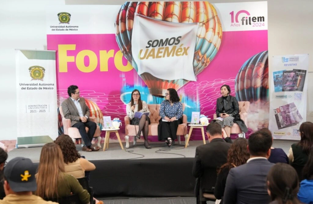 Panel de discusión en un foro con cuatro personas sentadas en sillones al frente del escenario. Sobre pequeñas mesas hay libros o revistas. Detrás se observa un fondo con diseño gráfico que incluye el texto “Somos UAEMéx” y elementos visuales llamativos, así como un banner con publicaciones. En primer plano se ven asistentes sentados escuchando la charla.