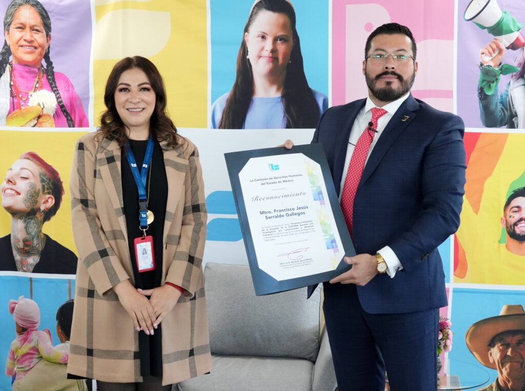 Dos personas posan de pie frente a un fondo colorido con imágenes de rostros y gráficos. Una de ellas sostiene un reconocimiento enmarcado con el texto visible “Reconocimiento” y el nombre de una persona. Detrás hay sillones y una decoración que sugiere un evento institucional de entrega de distinciones.