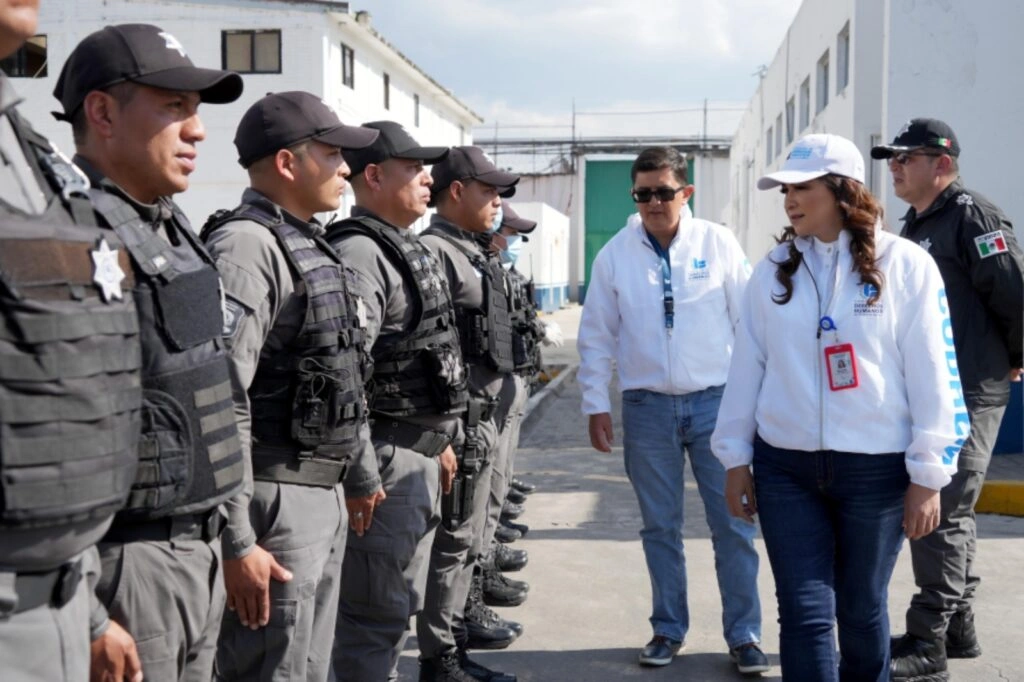 Grupo de elementos uniformados se encuentra formado en fila en un patio exterior de instalaciones institucionales con edificios blancos. Frente a personal operativo se observan dos personas con chamarras blancas con logotipos de una institución de derechos humanos, quienes recorren la fila dialogando. El entorno presenta piso de concreto, luz natural intensa y una puerta verde al fondo.
