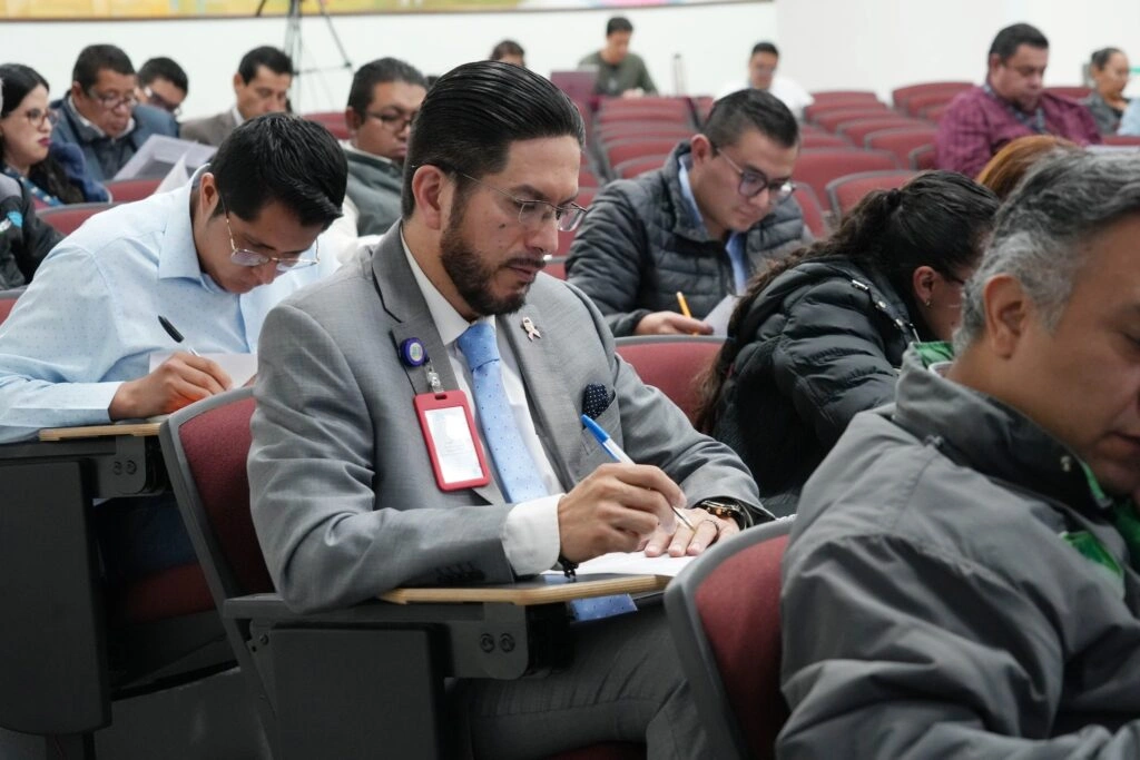 Asistentes a una capacitación sentados en butacas tipo auditorio tomando notas en libretas. En primer plano destaca una persona con traje formal escribiendo con pluma, mientras alrededor otras personas observan y participan de manera atenta. Se aprecia un ambiente de formación académica o institucional.