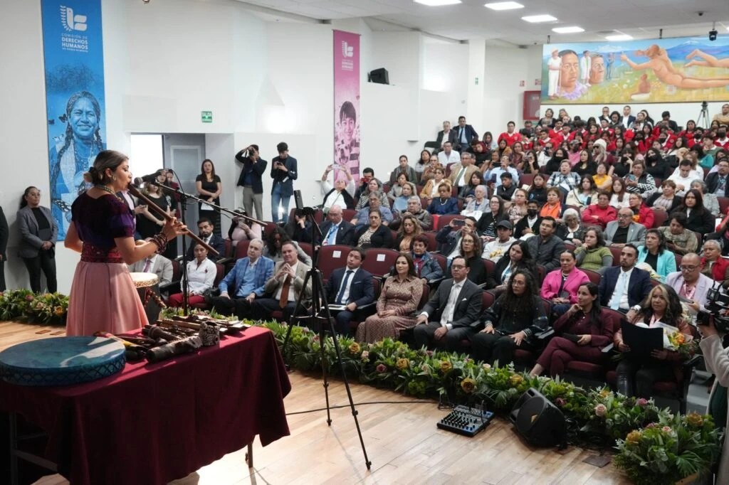 Auditorio con público numeroso sentado en gradas y butacas; al frente una persona habla en un micrófono junto a una mesa con instrumentos y objetos culturales. Hay fotógrafos y cámaras registrando el evento, con banners de la Comisión de Derechos Humanos en las paredes.