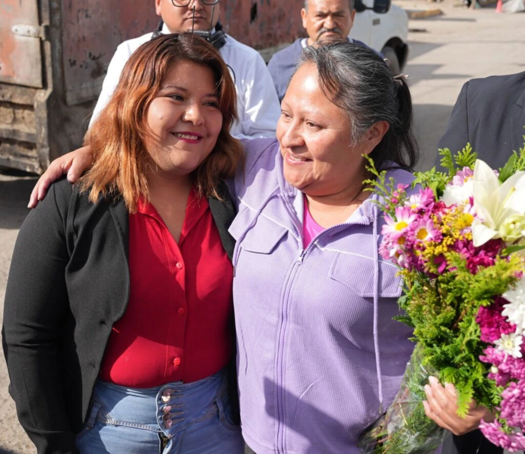 Dos personas de pie en un espacio exterior urbano, una de ellas sostiene un ramo de flores mientras ambas están con el brazo sobre el hombro de la otra; detrás se observan otras personas y una estructura metálica.