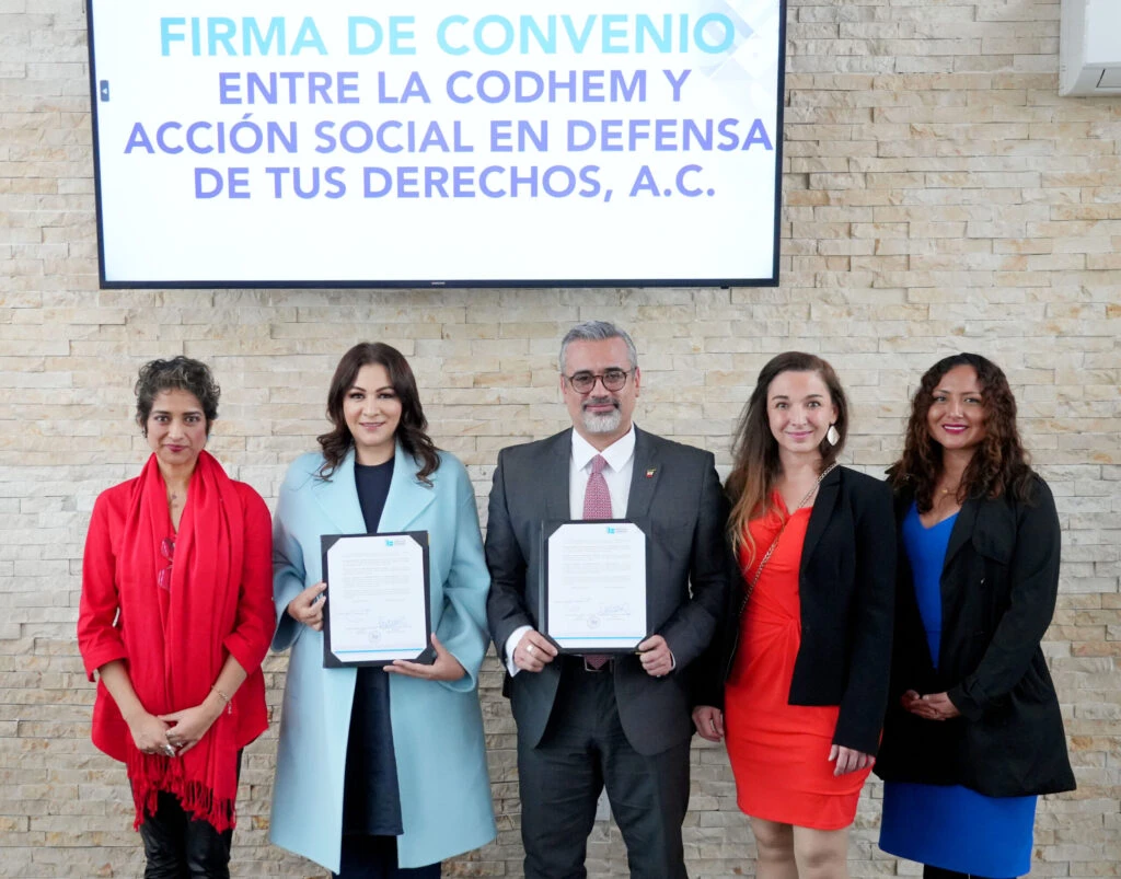 Grupo de cinco personas posando de pie frente a un muro de piedra, dos de ellas sostienen documentos firmados; arriba se observa una pantalla con el texto: “Firma de convenio entre la CODHEM y Acción Social en Defensa de tus Derechos, A.C.”.